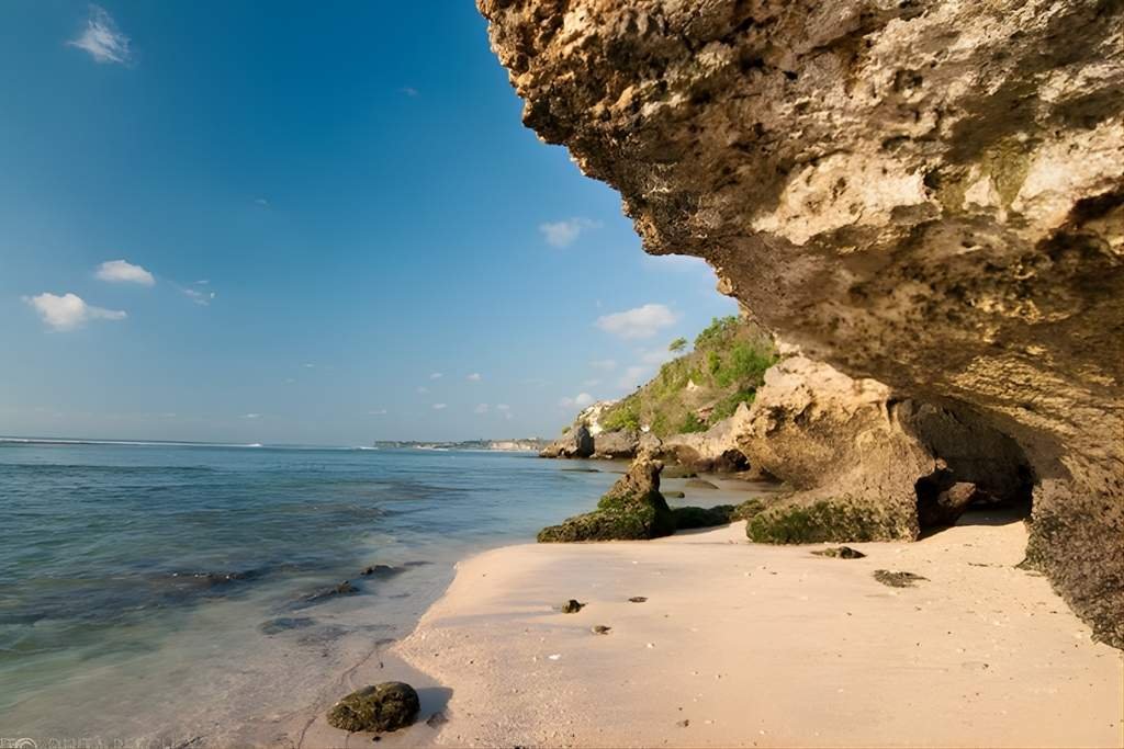 A quiet beach in Uluwatu, Bali, with soft sand, shallow clear water, and a large overhanging limestone cliff casting shade along the shore. The coastline curves into the distance under a bright blue sky.