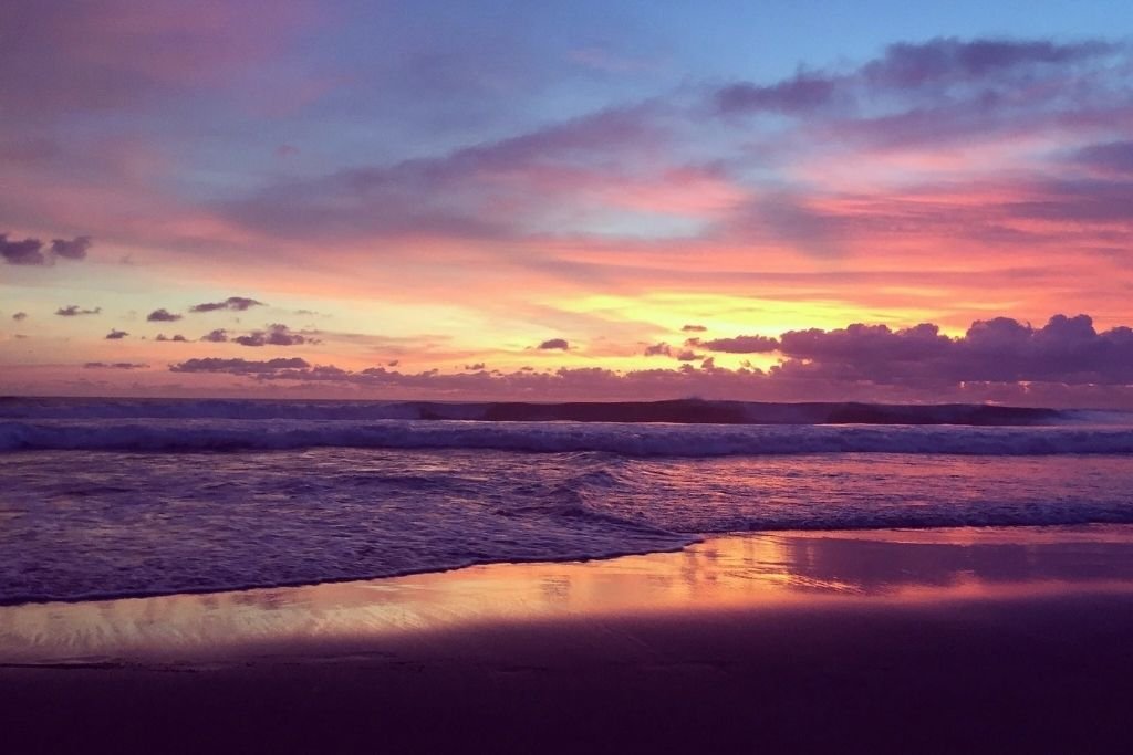 A vivid sunset over Echo Beach Bali, with colorful pink, purple, and orange hues reflecting on the wet sand and gentle waves rolling onto the shore.