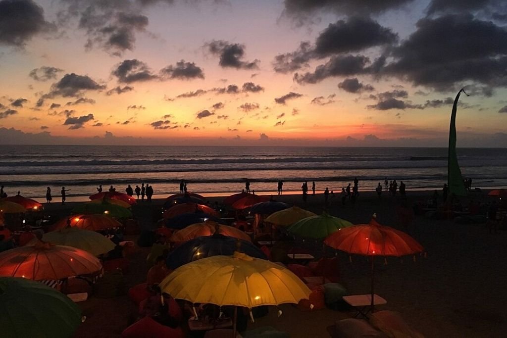 Evening view of Seminyak Beach in Bali, featuring colorful umbrellas and bean bags lit with soft lights, with silhouettes of people walking along the shoreline during a vibrant sunset, highlighting the allure of Seminyak and Petitenget.