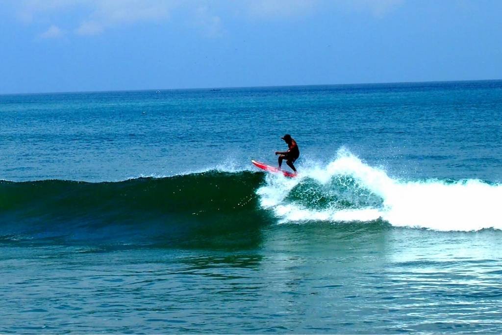 A surfer balances mid-ride on a pink surfboard, carving along a clean wave in the clear blue waters of Kuta Surf Bali under a bright sky.