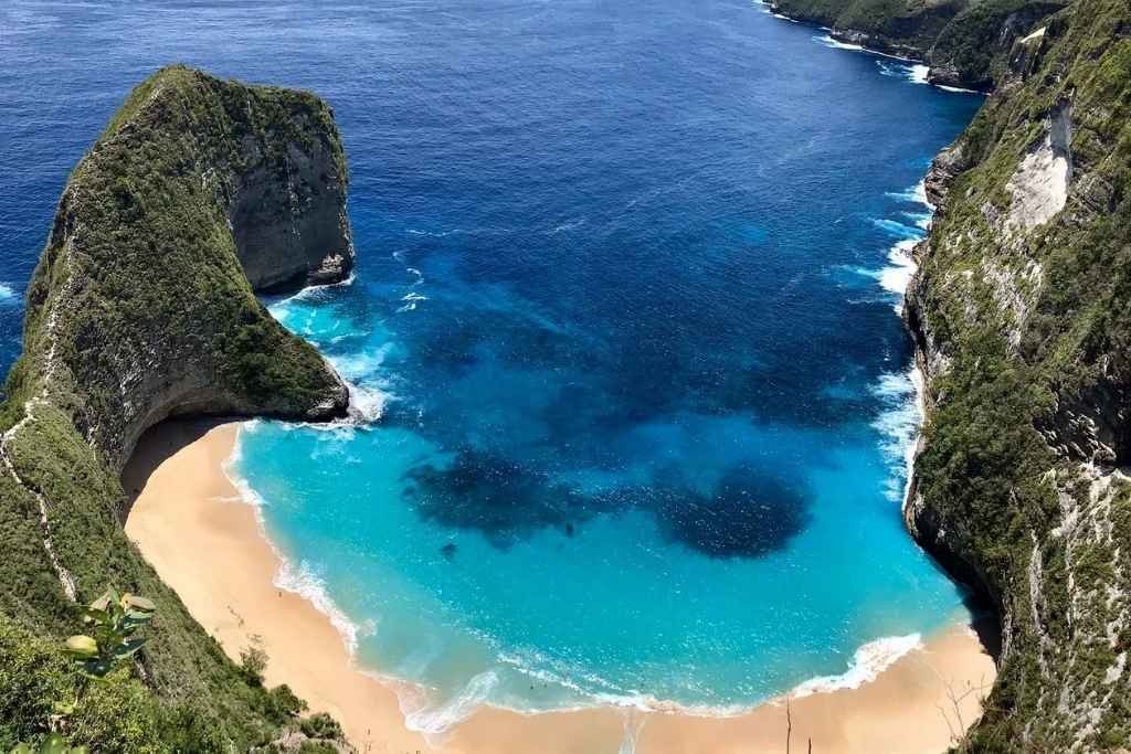 A breathtaking aerial view of Kelingking Beach on Nusa Penida Island, Bali, featuring turquoise waters, golden sand, and dramatic limestone cliffs resembling the shape of a T-Rex. The waves gently lap the shore in a secluded cove surrounded by lush greenery.