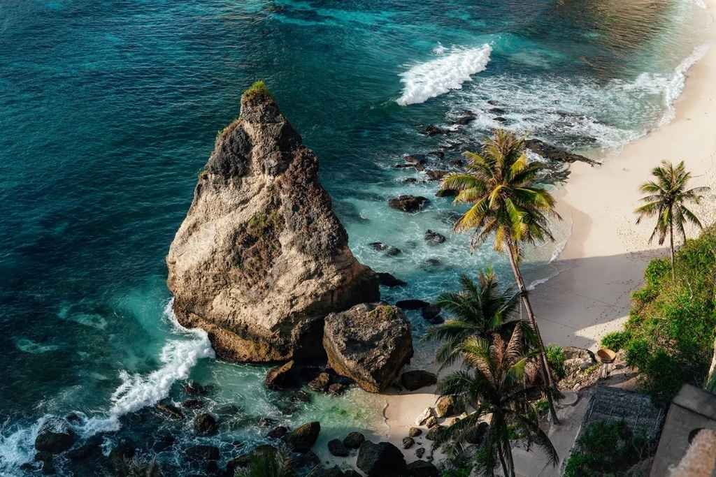 Aerial view of Padang Padang Beach in Bali with turquoise waters crashing gently against a small stretch of golden sand, surrounded by rocky cliffs and coral reef shadows beneath the surface.