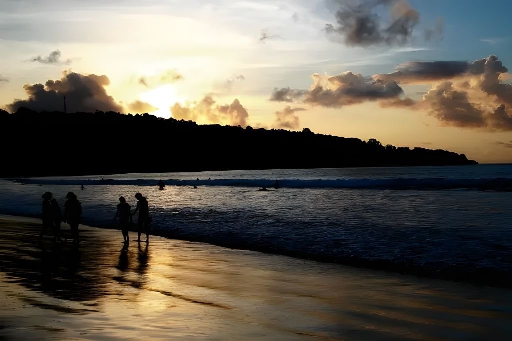 Silhouettes of people walking along the shoreline at Jimbaran Beach during sunset, with golden reflections on the wet sand and soft waves rolling in under a sky filled with glowing clouds.