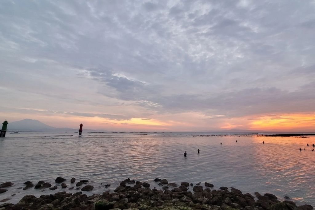 Tranquil Sanur coastline in Bali at sunset, with soft orange and pink hues reflecting off the calm ocean, silhouetted swimmers, and two lighthouse markers standing in the water with distant mountains in the background.