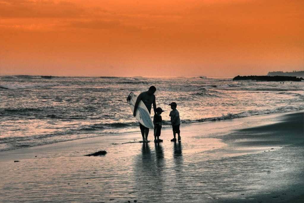 A surfer with two children standing on the shore of Canggu Beach, Bali, during sunset, with waves rolling in and the sky glowing orange.
