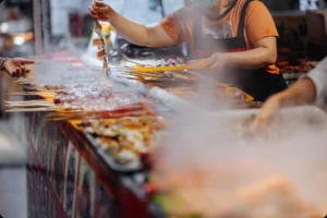 A busy street food vendor in Bali grills skewered seafood through rising steam, showcasing the vibrant flavors promoted by Coco Development Group as part of the best street food in Bali.