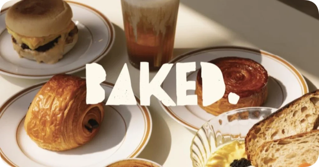 Home 213 Flat lay of artisanal breakfast items from Baked Bali, featuring a breakfast sandwich, pain au chocolat, cinnamon roll, crusty sourdough toast with spread, and a coffee, with the bold "BAKED." logo overlaid.