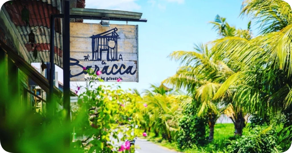 Home 211 Outdoor sign for La Baracca, a rustic Italian restaurant and pizzeria, hanging on a wooden board beside a tropical road lined with palm trees and blooming flowers.