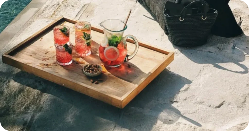 Wooden tray on a stone surface beside a pool, holding a pitcher of red fruit-infused drink with lemon slices and mint, three matching glasses, and a small bowl of almonds. The Lawn Canggu