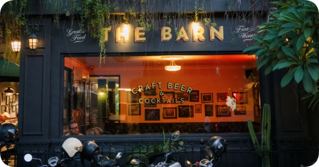 Home 209 Exterior of The Barn bar and restaurant with a dark facade, hanging greenery, and a large window displaying the text "Craft Beer & Cocktails" in gold lettering, with motorbikes parked out front.
