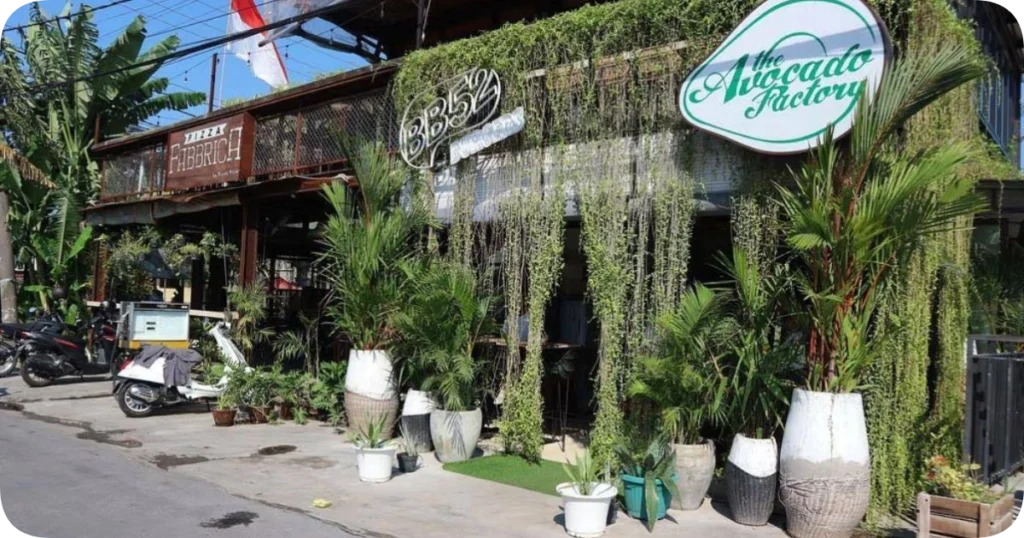 Exterior of The Avocado Factory, a lush, plant-covered restaurant with hanging greenery and large potted palms, next to Pizza Fabbrica canggu and BB52 Burgers on a sunny street lined with scooters.