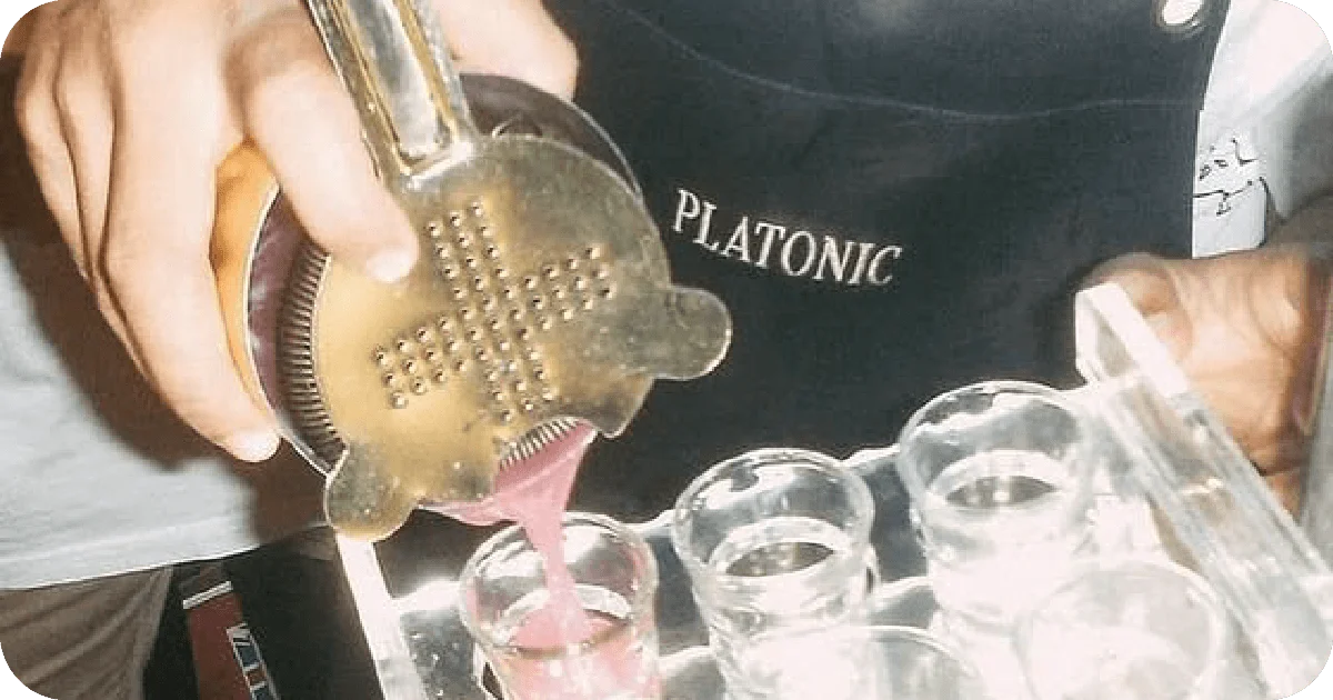 Bartender pouring pink shots into glasses, PLATONIC-branded apron