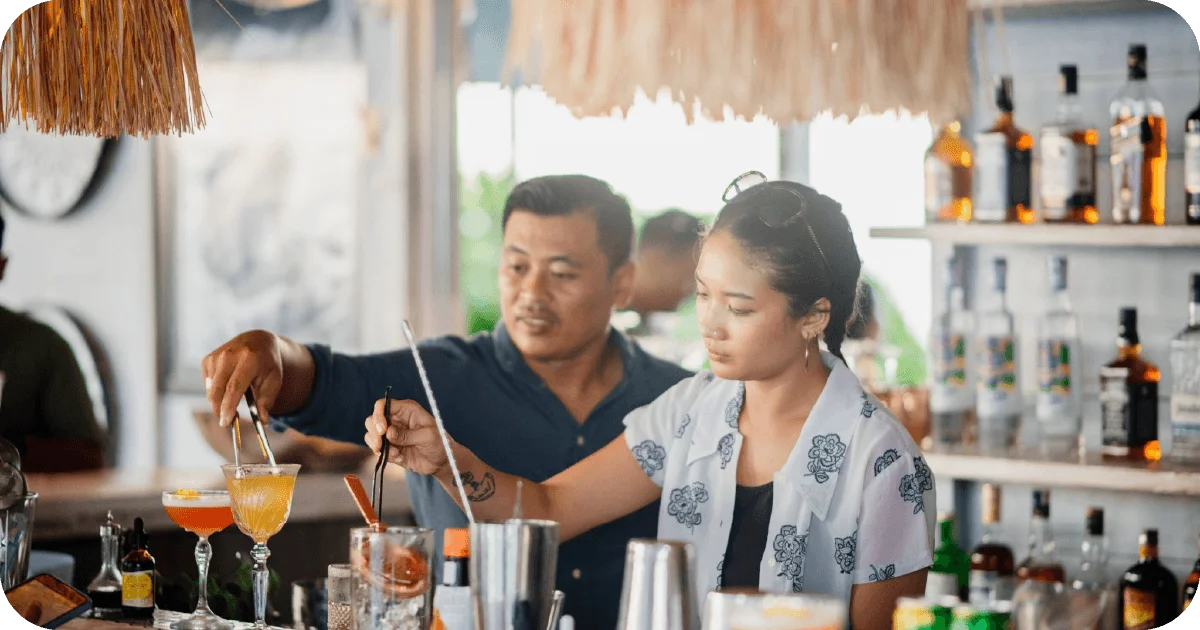 Bartenders mixing cocktails at backlit bar with spirits