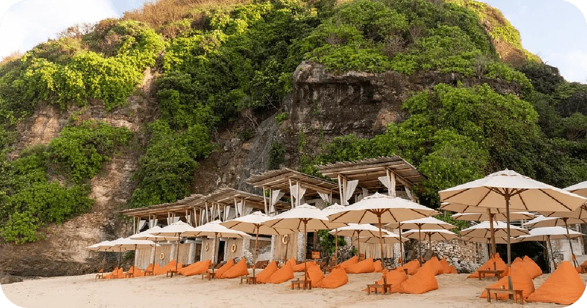Beachfront seating with white umbrellas and orange beanbags below cliffside greenery
