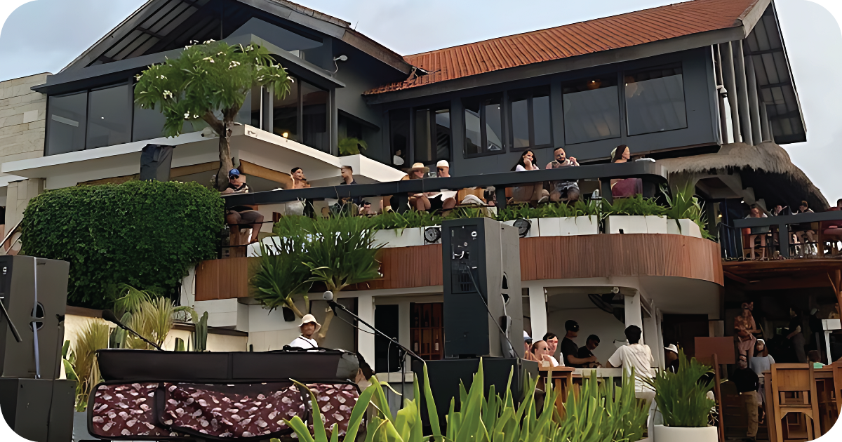 Multi-level beachfront restaurant with crowded balcony seating and speakers