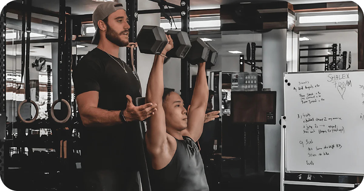 Personal training session with seated dumbbell press beside workout whiteboard