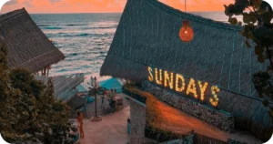 SUNDAYS illuminated sign overlooking ocean beside thatched roofs at sunset