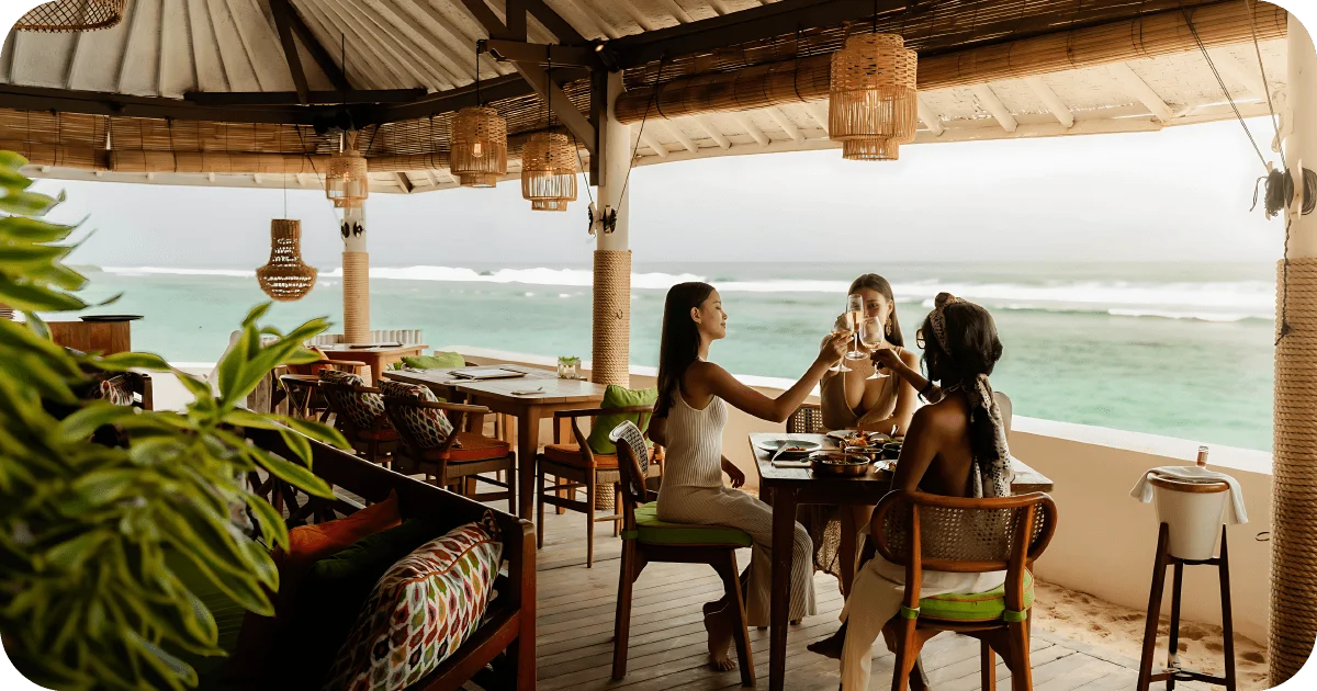Three guests toasting wine at oceanfront, rattan-lit beach bar