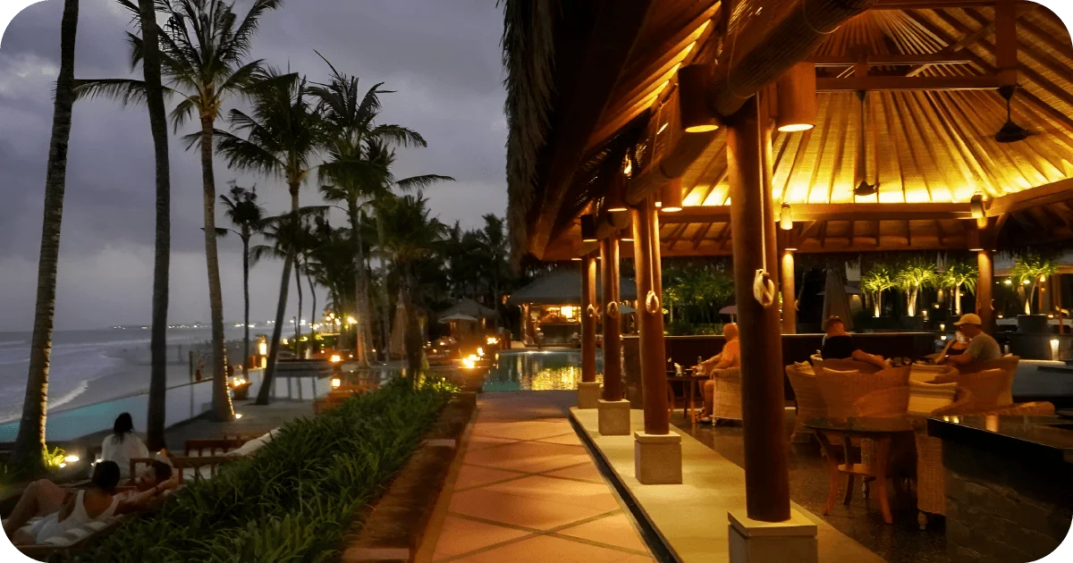 Beachfront poolside bar at dusk with palm trees and lanterns.