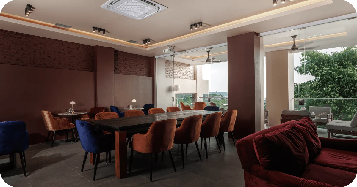 Bright lounge with long wood table, colorful chairs, and balcony greenery