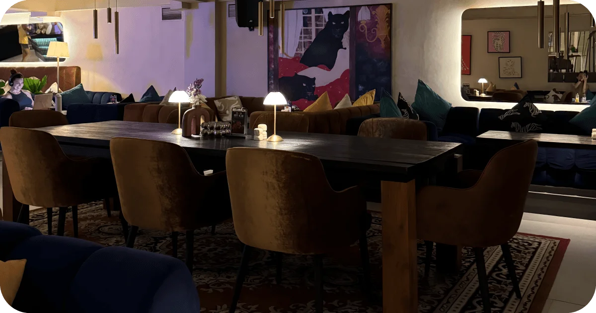 Dim lounge with long communal table, velvet chairs, and table lamps